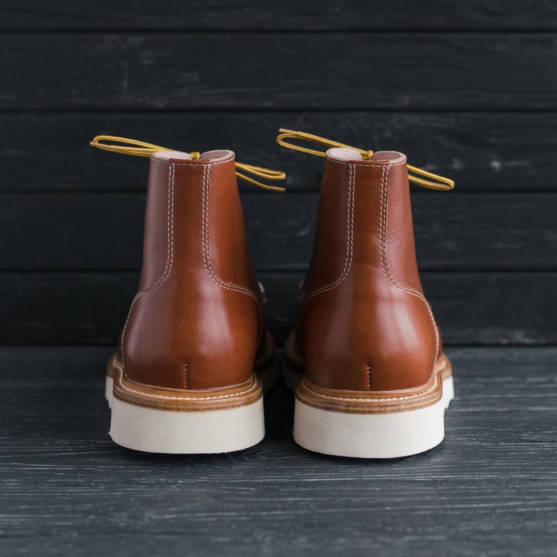 May include: A pair of brown leather boots with white soles. The boots have yellow laces and are shown from the back.
