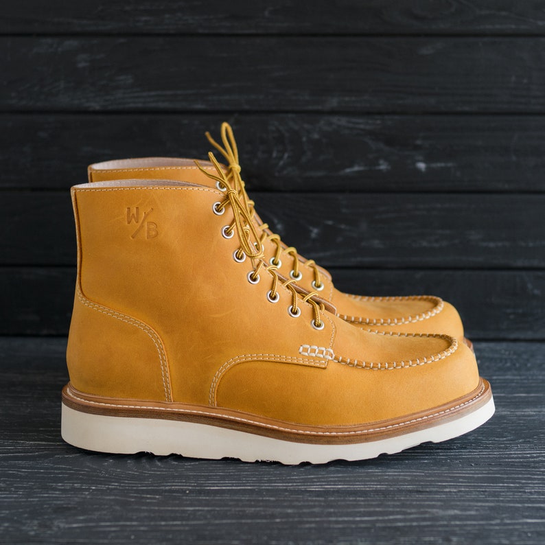 Puede incluir: Un par de botas de cuero marr&oacute;n con cordones y suela blanca. Las botas tienen un dise&ntilde;o de puntera mocas&iacute;n y el nombre de la marca "W/B" est&aacute; grabado en el lateral.