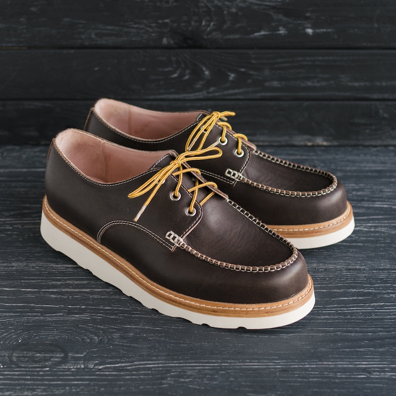 May include: A pair of brown leather shoes with white soles and yellow laces. The shoes have a moc toe design and a stitched seam around the top.