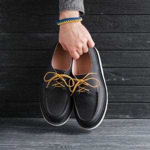 Peut inclure: Une paire de chaussures en cuir noir &agrave; lacets jaunes. Les chaussures sont tenues dans les mains d'une personne portant un bracelet bleu et jaune.