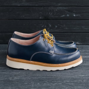 May include: A pair of blue leather shoes with a white sole and yellow laces. The shoes have a stitched detail around the toe and a slightly rounded toe shape.