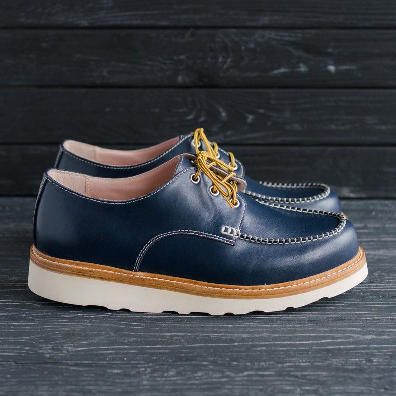 May include: A pair of blue leather shoes with a white sole and yellow laces. The shoes have a stitched detail around the toe and a slightly rounded toe shape.