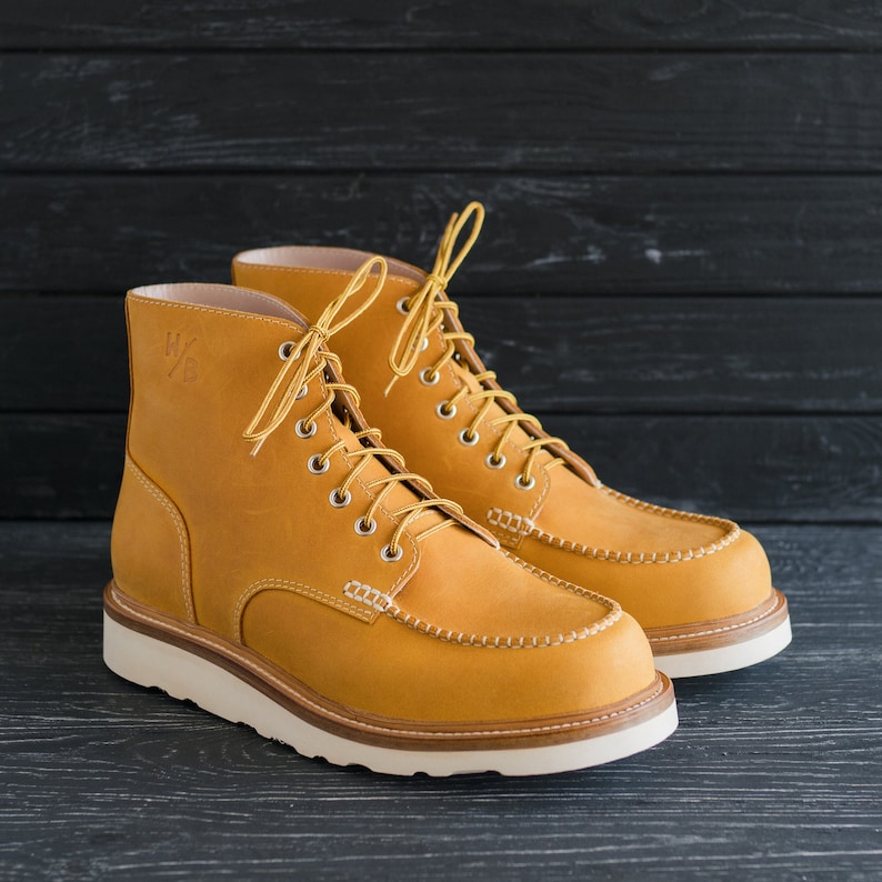 Puede incluir: Un par de botas de cuero amarillo con cordones blancos y suela blanca. Las botas tienen un dise&ntilde;o de punta mocas&iacute;n y est&aacute;n hechas de cuero suave y flexible.