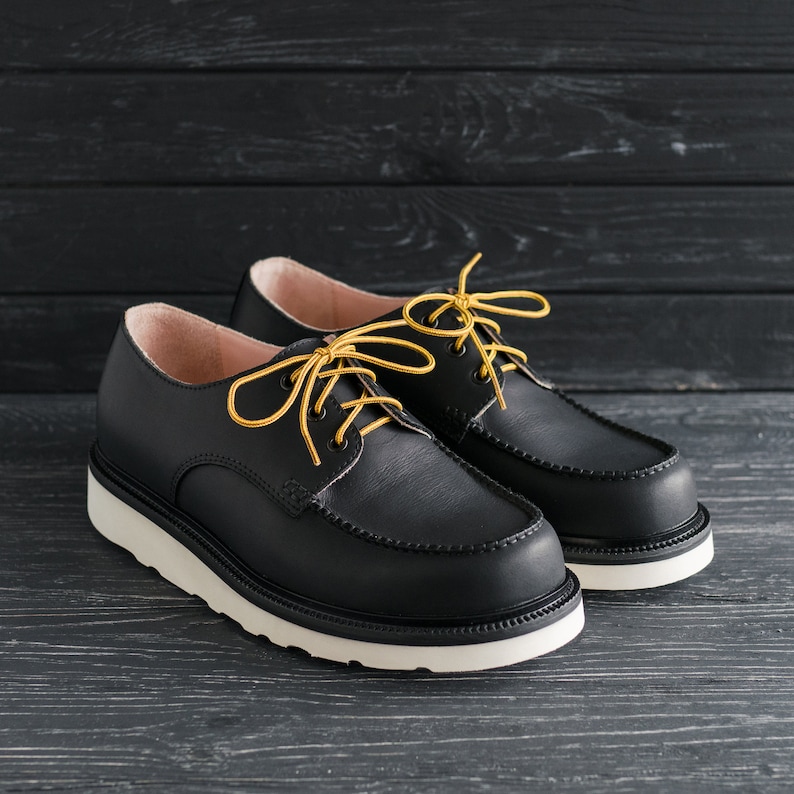 Peut inclure: Deux chaussures en cuir noir avec des semelles blanches et des lacets jaunes. Les chaussures ont un style d&eacute;contract&eacute; et confortable.