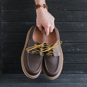 May include: A pair of brown leather boat shoes with yellow laces. The shoes are being held in someone's hand.