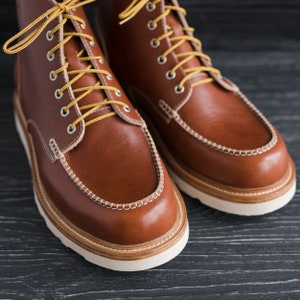 May include: A pair of brown leather boots with yellow laces. The boots have a moc toe design and a white stitching detail around the top of the boot.