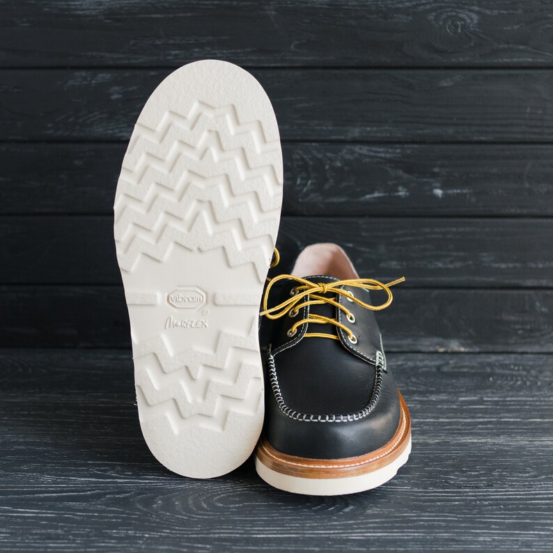 May include: A pair of black leather shoes with yellow laces. The shoes have a white rubber sole with a textured pattern. The sole is labeled "Vibram" and "Margex".