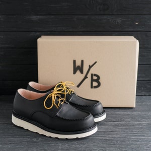 Peut inclure: Une paire de chaussures en cuir noir &agrave; lacets avec une semelle blanche et des lacets jaunes. Les chaussures sont pos&eacute;es sur une surface en bois noir devant une bo&icirc;te en carton sur laquelle est imprim&eacute; le texte "W/B".
