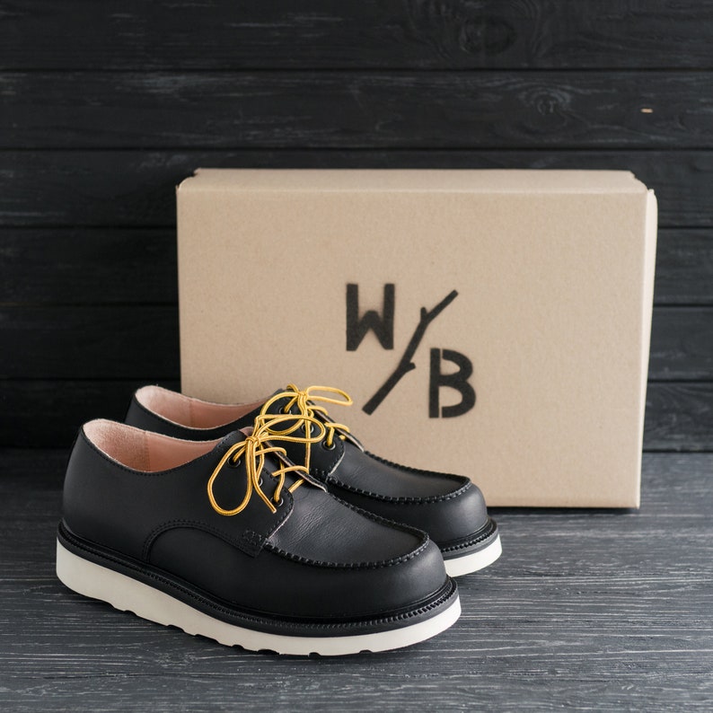 Peut inclure: Une paire de chaussures en cuir noir &agrave; lacets avec une semelle blanche et des lacets jaunes. Les chaussures sont pos&eacute;es sur une surface en bois noir devant une bo&icirc;te en carton sur laquelle est imprim&eacute; le texte "W/B".