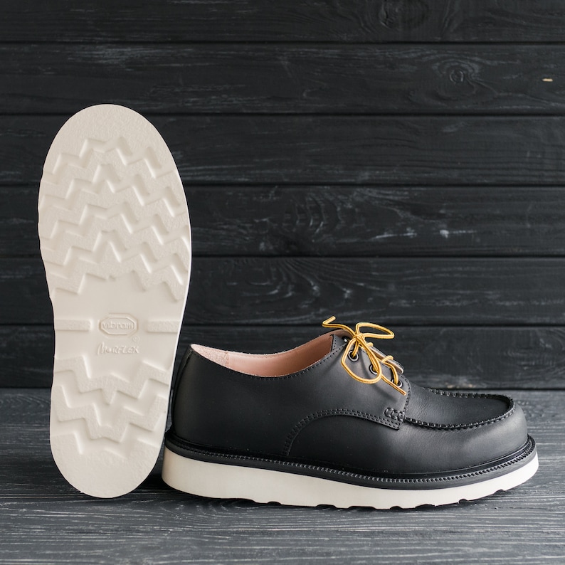 Peut inclure: Une paire de chaussures en cuir noir avec des semelles blanches et des lacets jaunes. Les chaussures ont une pointe arrondie et un talon bas. Les chaussures sont pos&eacute;es sur une surface en bois noir.