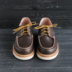 May include: A pair of brown leather boat shoes with yellow laces. The shoes have a white rubber sole and a brown leather upper.