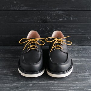 Peut inclure: Une paire de chaussures en cuir noir &agrave; lacets jaunes. Les chaussures ont une semelle blanche et sont sur un fond en bois noir.