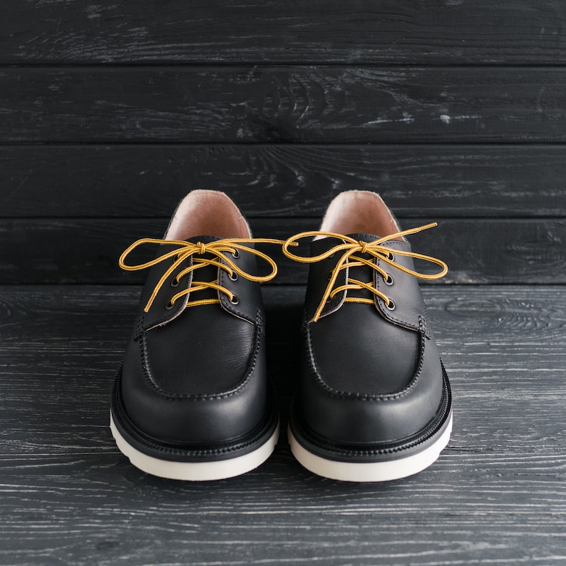 Peut inclure: Une paire de chaussures en cuir noir &agrave; lacets jaunes. Les chaussures ont une semelle blanche et sont sur un fond en bois noir.