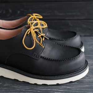Peut inclure: Une paire de chaussures en cuir noir &agrave; lacets jaunes. Les chaussures ont une semelle en caoutchouc blanche et une pointe en mocassin.