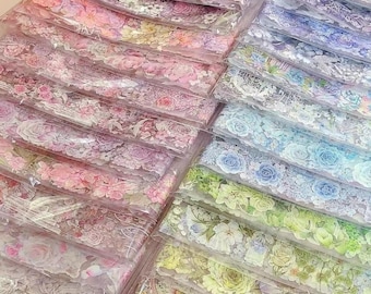 May include: An assortment of floral decorative tapes in various colors, including pink, purple, blue, and green. Each tape is packaged in a clear plastic sleeve, showcasing the intricate flower designs. The tapes feature roses, hydrangeas, and other floral patterns.