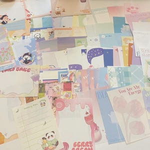 May include: A collection of colorful stationery items, including notepads, cards, and sticky notes. The designs feature illustrations of bears, pandas, and desserts, with text like "STRO CAKE" and "You Are My Energy". The color palette includes pastels.