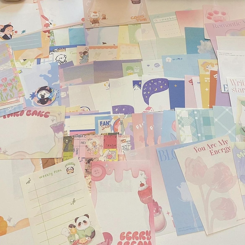 May include: A collection of colorful stationery items, including notepads, cards, and sticky notes. The designs feature illustrations of bears, pandas, and desserts, with text like "STRO CAKE" and "You Are My Energy". The color palette includes pastels.