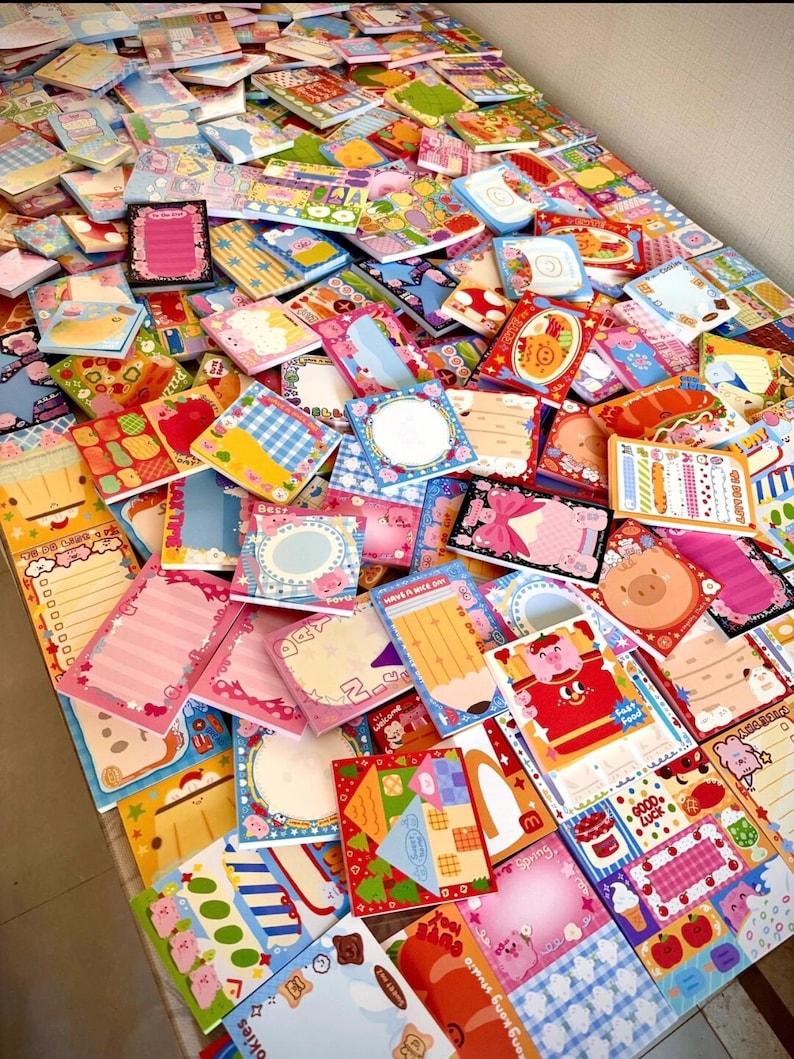 May include: A large assortment of colorful, decorative notepads with various designs, including pigs, food, and patterns. The notepads are in various shapes and sizes, with blank spaces for writing. The image is a top-down view of the notepads.