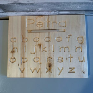 May include: A wooden alphabet board with the name "Petra" carved at the top. The lowercase alphabet is carved below the name. The board is made of light-colored wood.