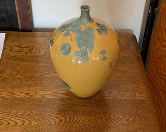 Puede incluir: Un jarrón de cerámica amarillo con cuerpo redondo y cuello estrecho. El jarrón está decorado con motivos florales azules y verdes.