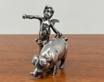 May include: A silver figurine of a cherub riding a pig. The cherub has wings and is holding onto the pig's back. The pig is walking forward with its head down.