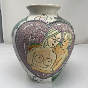 May include: A ceramic vase with a heart-shaped design. The vase features an illustration of two figures embracing, with a textured, speckled background. The colour palette includes cream, purple, green, and tan. The vase has a wide body and a narrow neck.