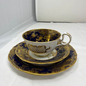 May include: A classic tea set comprising a teacup, saucer, and side plate. The set displays a navy blue base adorned with gold floral and lattice designs. The teacup is white with a gold handle, a timeless design.