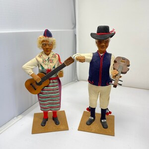 Könnte beinhalten: Zwei antike Volksfiguren in traditioneller Kleidung. Die Figur links hält eine Gitarre und die Figur rechts hält eine Geige. Beide Figuren stehen auf Holzsockeln.