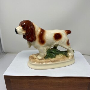 May include: A ceramic figurine of a brown and white dog with floppy ears standing on a base with green leaves.