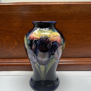 Puede incluir: Un jarrón de cerámica azul oscuro con un motivo floral pintado a mano con pétalos amarillos, rosas y verdes. El jarrón tiene un cuerpo redondeado, un cuello estrecho y un borde ensanchado, con un acabado brillante y pinceladas visibles.