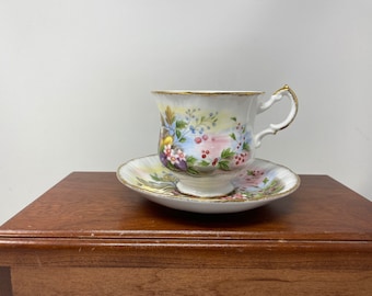 May include: A white teacup and saucer with a floral pattern in shades of pink, blue, yellow, and green. The cup has a gold rim and a delicate handle. The saucer has a scalloped edge.