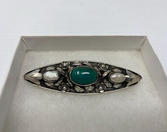 May include: A silver-toned brooch with an oval, dark green center stone and two off-white, oval-shaped pearls. The brooch is decorated with floral and leaf details.