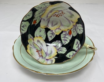May include: A vintage teacup and saucer set with a black background and white floral design. The saucer is a pale green color with a gold trim.