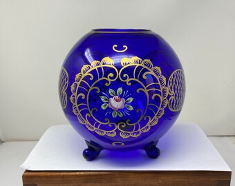 Op de afbeelding: Een kobaltblauwe glazen vaas met gouden geschilderde bloemenmotieven en drie poten. De vaas staat op een houten oppervlak.