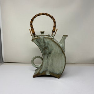 May include: A ceramic teapot with a unique, abstract shape and a light green glaze. The teapot has a brown woven handle and a spout on the side.