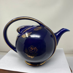 Peut inclure: Une théière bleu cobalt avec une poignée dorée et des motifs floraux dorés. La théière a un corps rond et un petit bec.