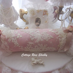 Könnte beinhalten: Ein rosa, zylindrisches Zierkissen mit weißer Spitzenauflage und floralen Akzenten. Das Kissen ist mit rosa Bändern und Rosen verziert. Der Text "Cottage Rose Shabby" ist sichtbar.
