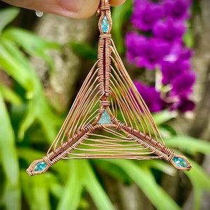 Può includere: Un ciondolo triangolare avvolto in filo di rame con tre pietre preziose blu chiaro. Il ciondolo è appeso a un anello di filo di rame.