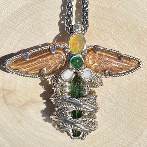 May include: A silver wire-wrapped pendant featuring a green tourmaline crystal, a golden sunstone, and an opal. The pendant is shaped like an angel with wings made of sunstone.