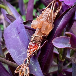 Puede incluir: Un colgante envuelto en alambre de cobre con un grupo de cristales de selenita naranja y un cristal rojo más pequeño. El colgante se muestra sobre un fondo de follaje morado.