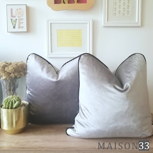 May include: Two gray velvet throw pillows with black piping. The pillows are on a wooden surface.