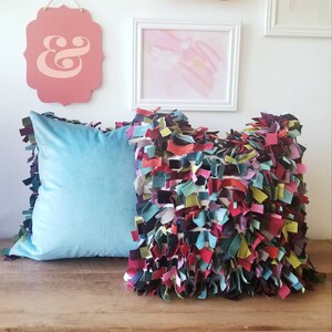 May include: Two decorative pillows, one is a solid light blue velvet pillow with a fringe border, the other is a multi-colored pillow with a fringe border made of fabric scraps in shades of red, pink, purple, green, blue, and white.