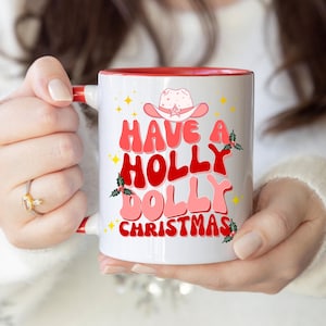 May include: White ceramic mug with a red interior and handle, featuring the text "Have a Holly Dolly Christmas" in pink and red, with a cowboy hat graphic. The mug is decorated with holly and stars.
