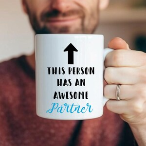 May include: White ceramic mug with a black arrow pointing upwards and the text "This person has an awesome partner" in black lettering. The word "partner" is in blue lettering.