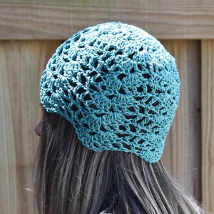 May include: A teal crocheted hat with an open, lacy design. The hat is worn on a person's head, with the back of the head and long, light brown hair visible. The hat's texture and color are the focal points, set against a wooden fence background.