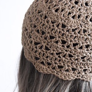 Puede incluir: Un gorro de ganchillo marr&oacute;n con un intrincado patr&oacute;n de estrellas caladas repetitivas. La prenda est&aacute; confeccionada con hilo de textura visible y presenta un borde festoneado en el dobladillo inferior que descansa sobre cabello oscuro.
