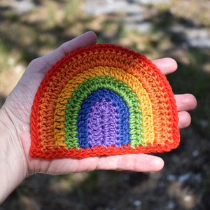 May include: A crocheted rainbow made with different colored yarn. The rainbow is in the shape of a half circle and is held in a person's hand.