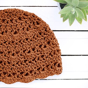 Puede incluir: Un gorro de color &oacute;xido que presenta un patr&oacute;n de ganchillo calado repetitivo. El gorro est&aacute; confeccionado con hilo grueso con un dise&ntilde;o de borde festoneado, mostrando una estructura de puntos texturizada e interconectada en la prenda.