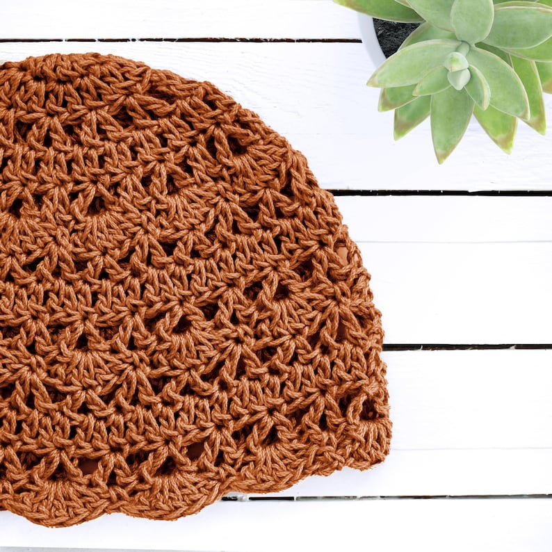 Puede incluir: Un gorro de color &oacute;xido que presenta un patr&oacute;n de ganchillo calado repetitivo. El gorro est&aacute; confeccionado con hilo grueso con un dise&ntilde;o de borde festoneado, mostrando una estructura de puntos texturizada e interconectada en la prenda.