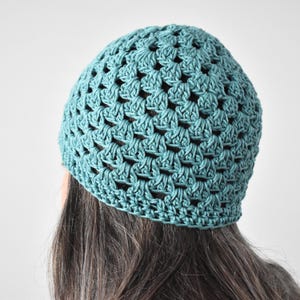May include: A teal blue crochet beanie featuring a classic granny square stitch pattern. The hat is constructed with a repeating openwork design that creates a textured, geometric appearance across the entire surface of the rounded head covering.